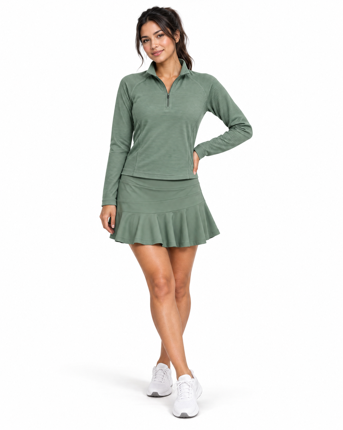 A woman modeling a matching sage green athletic set consisting of a long-sleeve quarter-zip performance top and a ruffled mini skort, paired with white sneakers, against a plain white studio background.