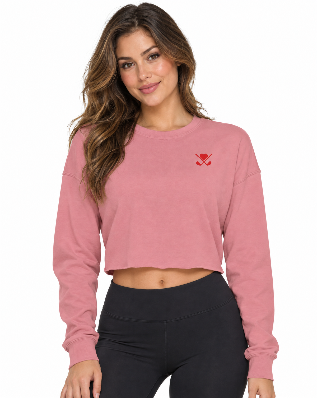 A woman modeling a dusty pink cropped long-sleeve sweatshirt with a small red embroidered logo on the left chest, paired with black leggings, against a plain white background.