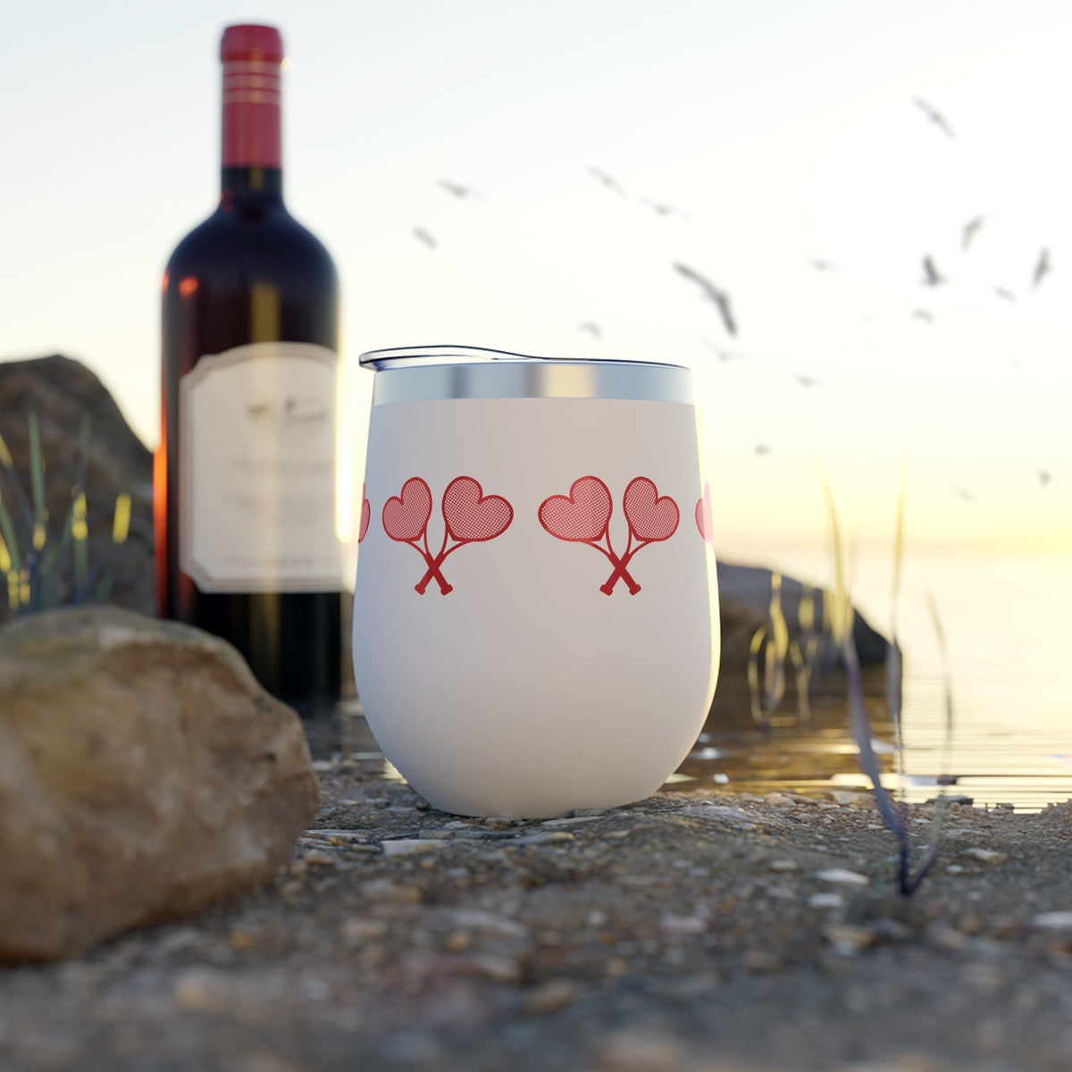 White tumbler with red heart designs on a beach with a bottle of wine