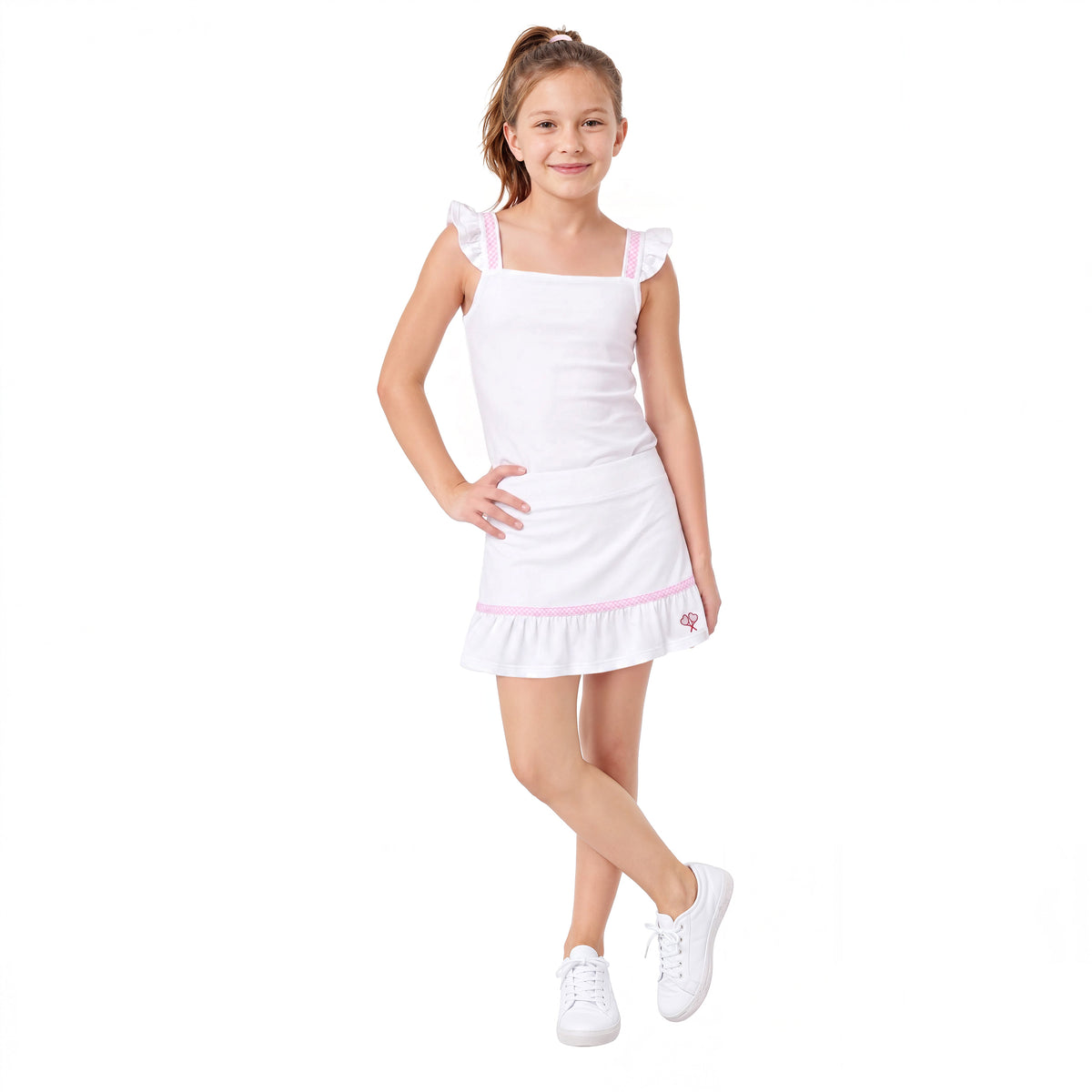 A young girl modeling a white sleeveless tennis top and skirt with ruffled shoulder straps and a hem, featuring a small pink logo on the bottom right. She is wearing white sneakers against a solid white background.
