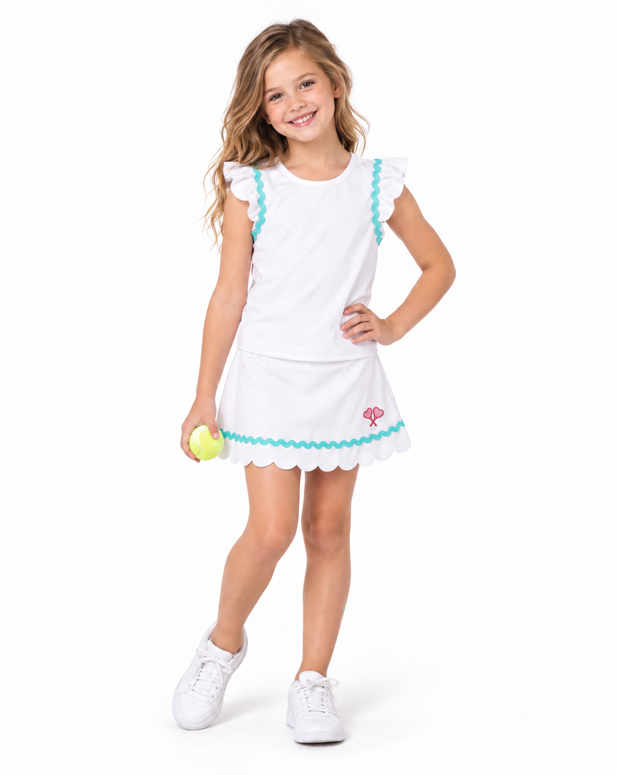 A young girl with blonde hair stands against a white studio background wearing a white tennis-style dress with turquoise trim and a small red tennis racket graphic near the hem. She is holding a yellow tennis ball and wearing white sneakers.