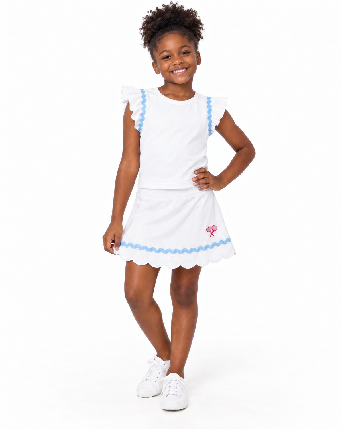 A young girl modeling a white two-piece outfit consisting of a short-sleeve top and a matching flared skirt, both decorated with light blue scalloped trim. The skirt features a small pink embroidered flower on the lower left side.
