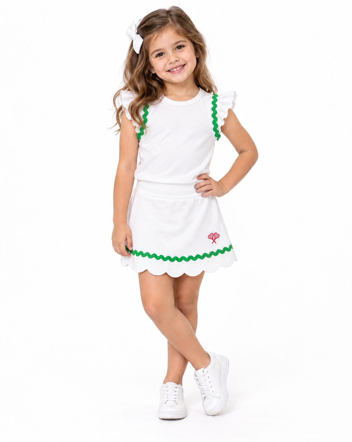 A young girl with long brown hair wearing a white sleeveless dress with green scalloped trim and a small red logo, styled with white sneakers and a white hair bow, posing against a solid white background.