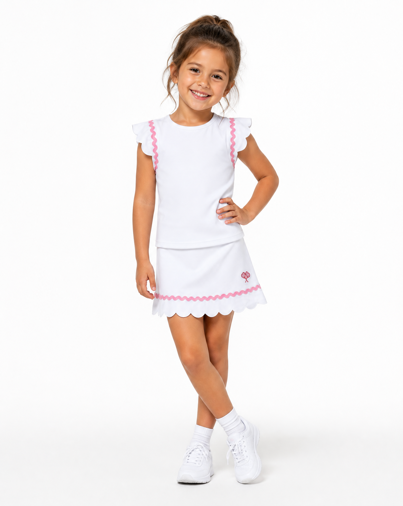 A young girl modeling a white two-piece dress set with pink scalloped trim details, consisting of a short-sleeve top and a matching skirt, paired with white socks and sneakers against a plain white background.