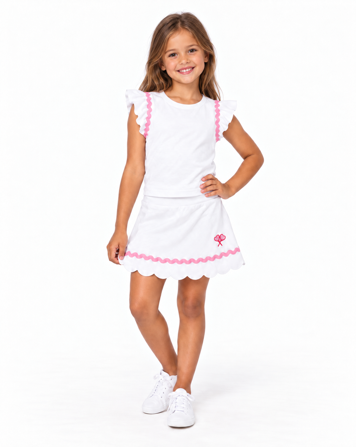A young girl modeling a white two-piece outfit consisting of a short-sleeve top with pink ruffle trim and a matching skirt with a scalloped hem and pink bow detail, paired with white sneakers on a white studio background.