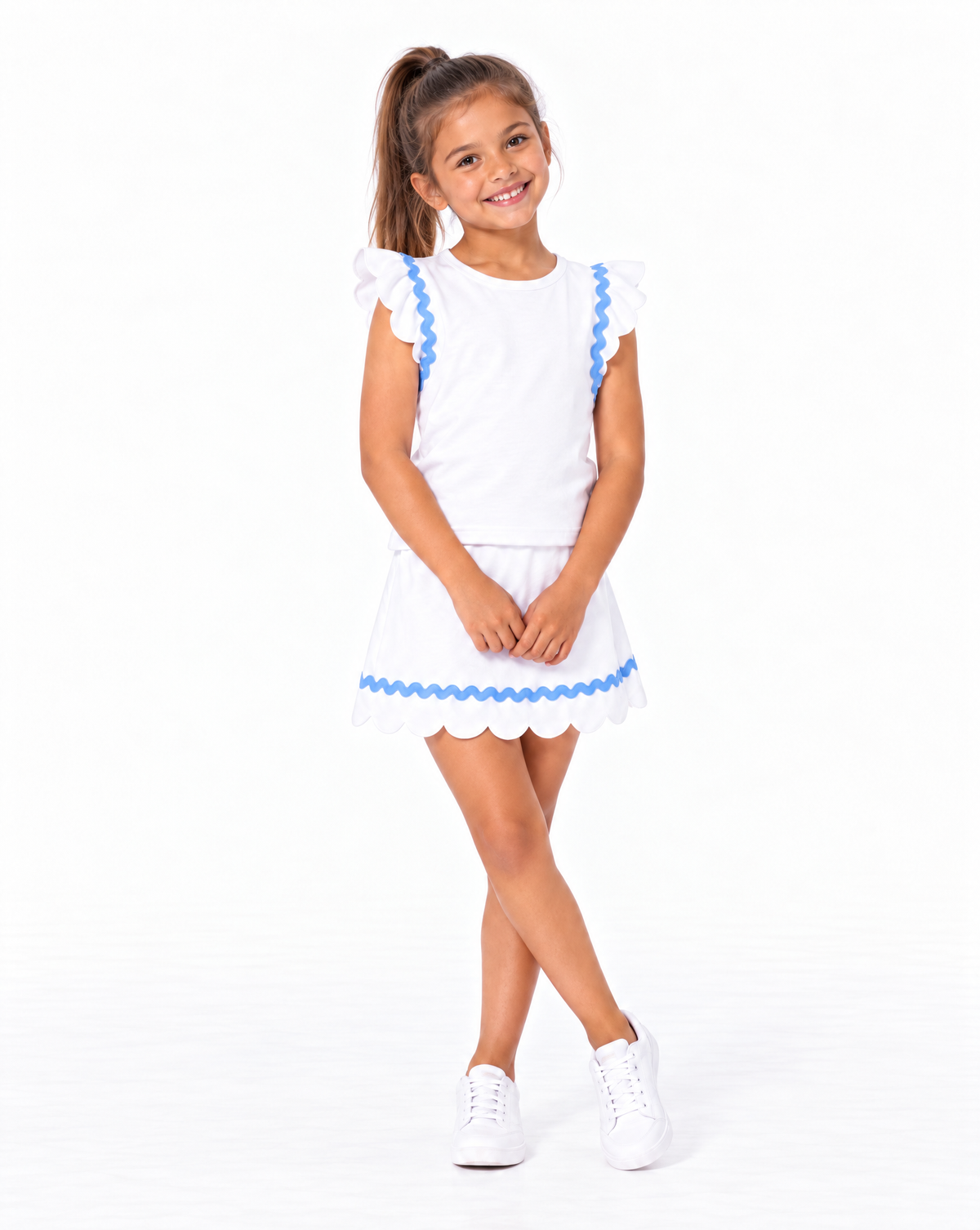 A young girl with a high ponytail is modeling a white sleeveless top and skirt with blue scalloped trim, paired with white sneakers, against a solid white studio background.