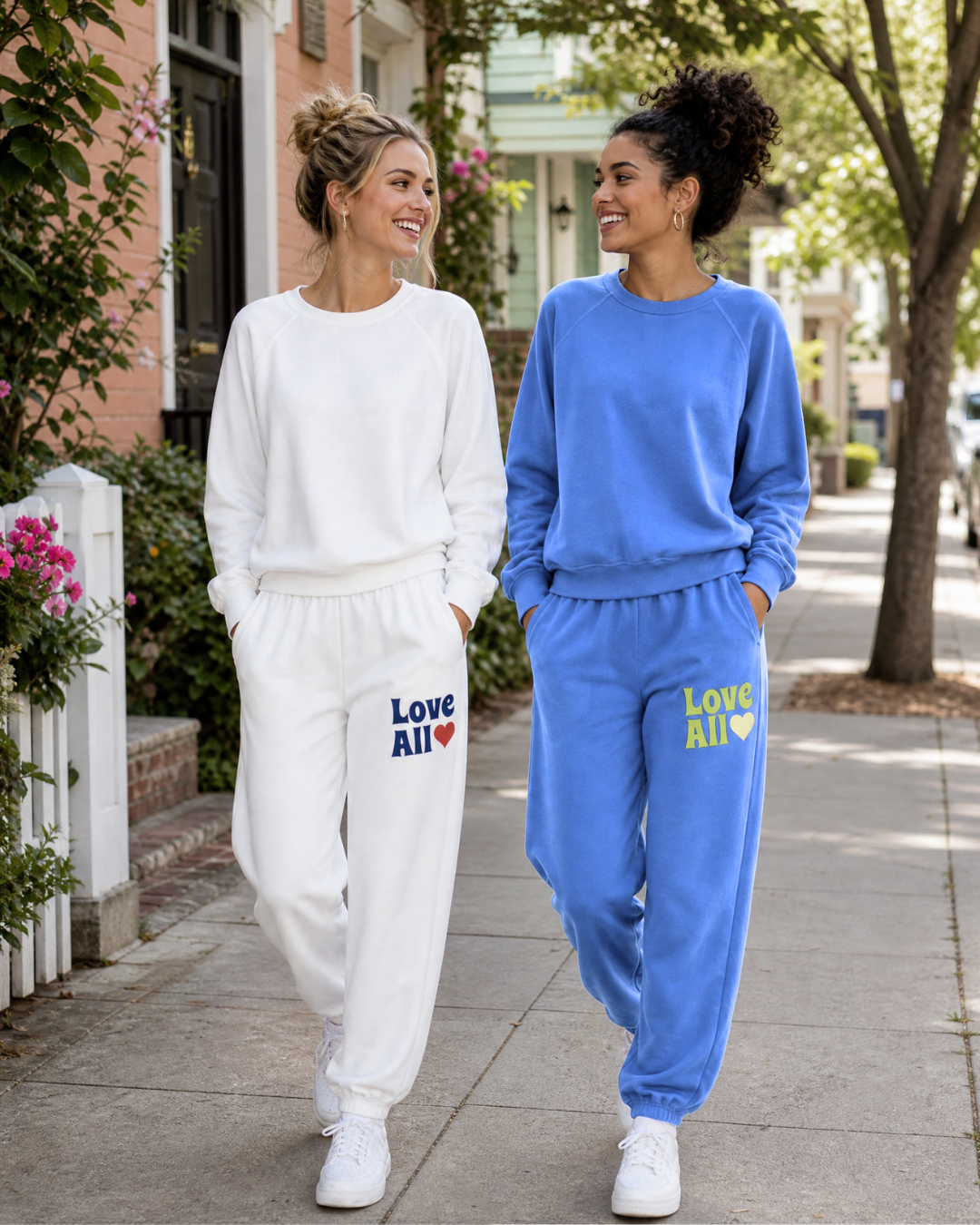 Two women walking side-by-side on a sidewalk wearing matching sweatshirt and jogger sets. The woman on the left wears a white set with 'Love All' printed in blue and red on the left leg.