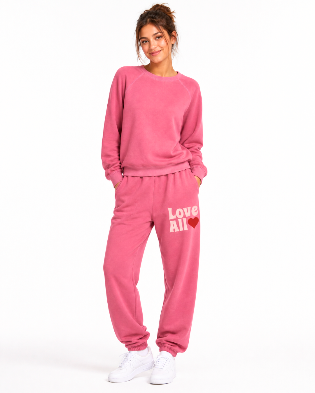 A woman modeling a matching pink sweatshirt and jogger pants set with white sneakers, standing against a plain white studio background. The sweatshirt has a crew neck and long sleeves, while the joggers feature a white 'Love All' text and red heart graphi