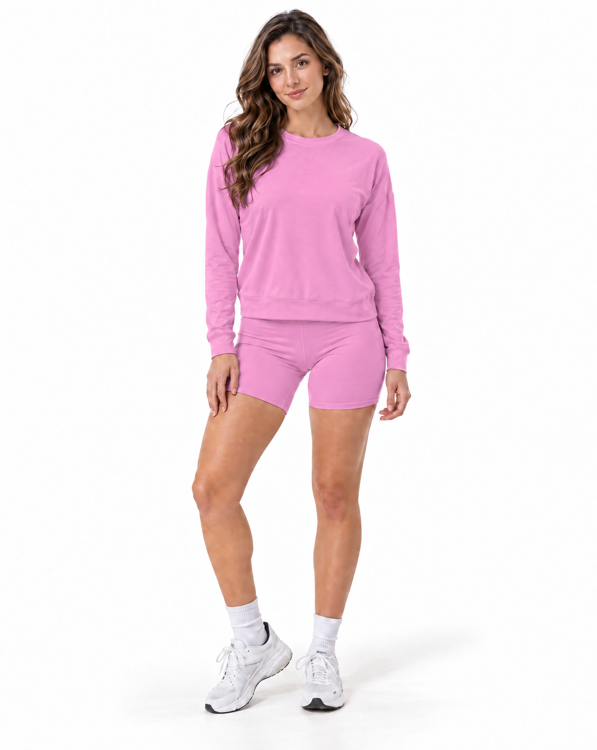 A woman modeling a matching pink activewear set consisting of a long-sleeve crewneck sweatshirt and matching athletic shorts, paired with white socks and white sneakers, against a plain white background.