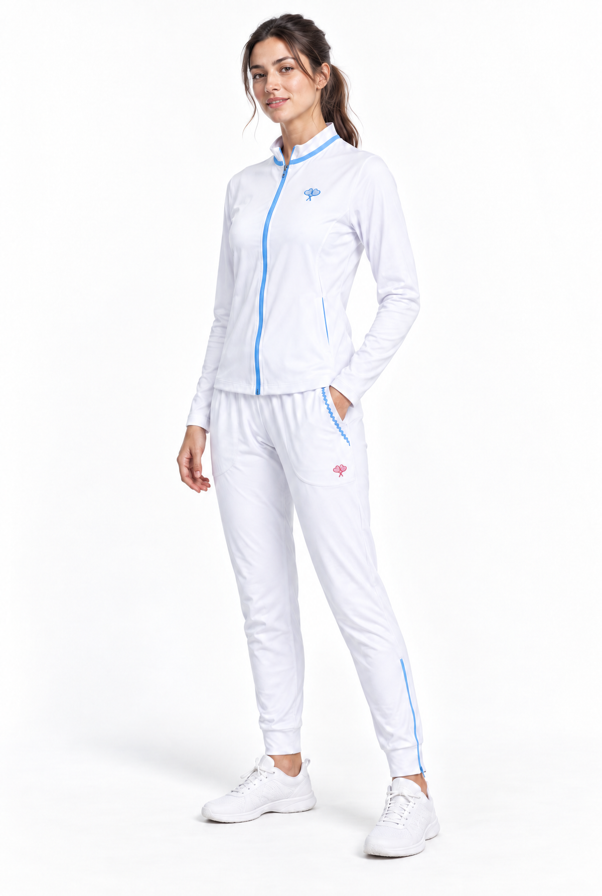 A woman modeling a white athletic tracksuit with light blue zipper accents and small pink logo details, paired with white sneakers, against a plain white studio background.