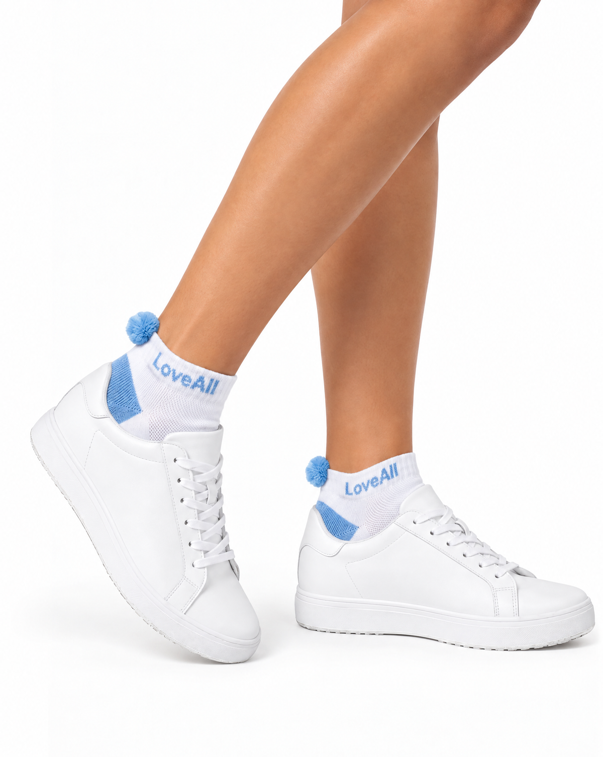 White low-top sneakers worn with white socks featuring blue pom-poms and the brand name 'LoveAll' printed in blue, shown on a model's feet against a white background.