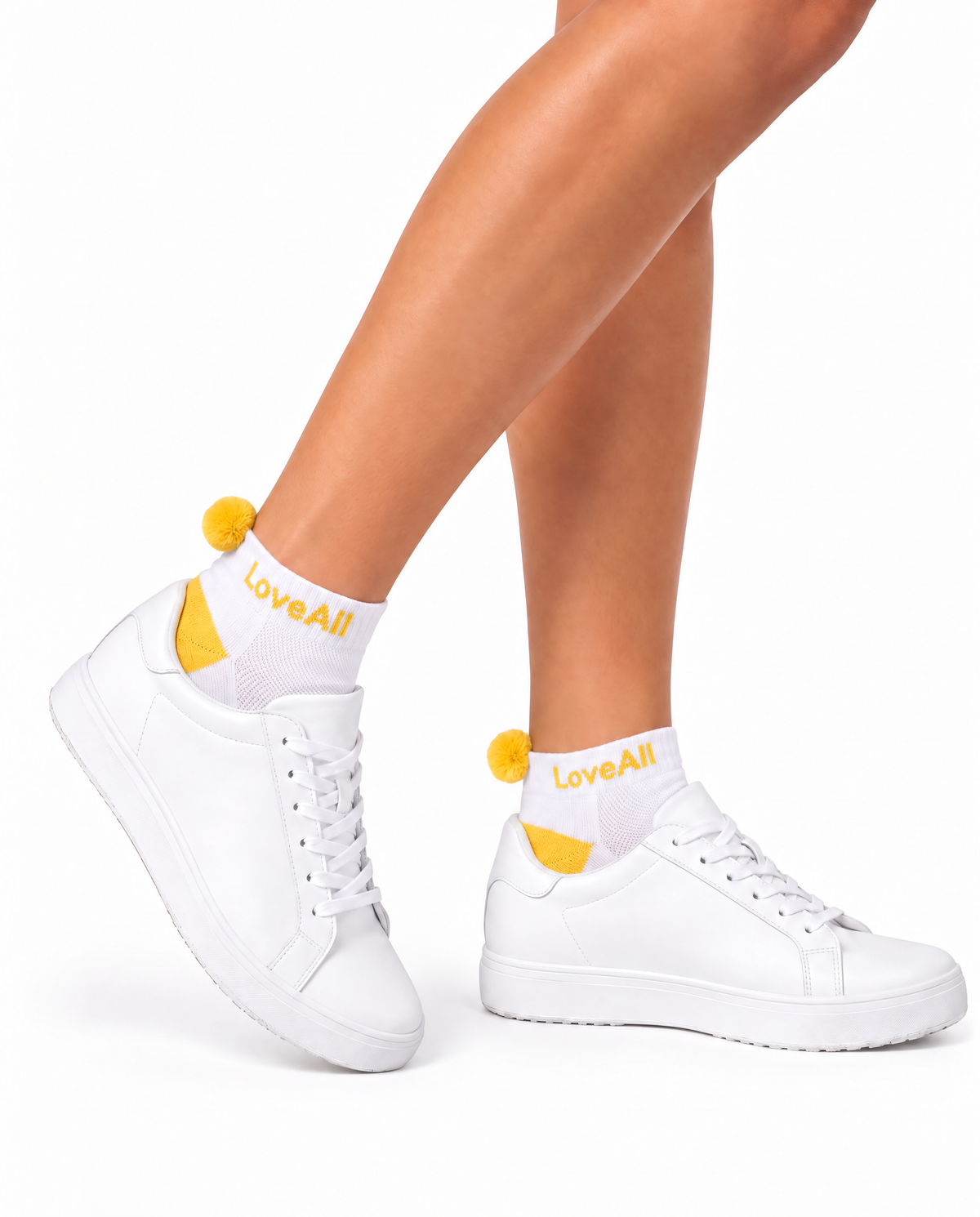 White low-top sneakers worn with white crew socks featuring yellow pom-poms and the brand name 'LoveAll' printed in yellow, shown on a model's feet against a white background.