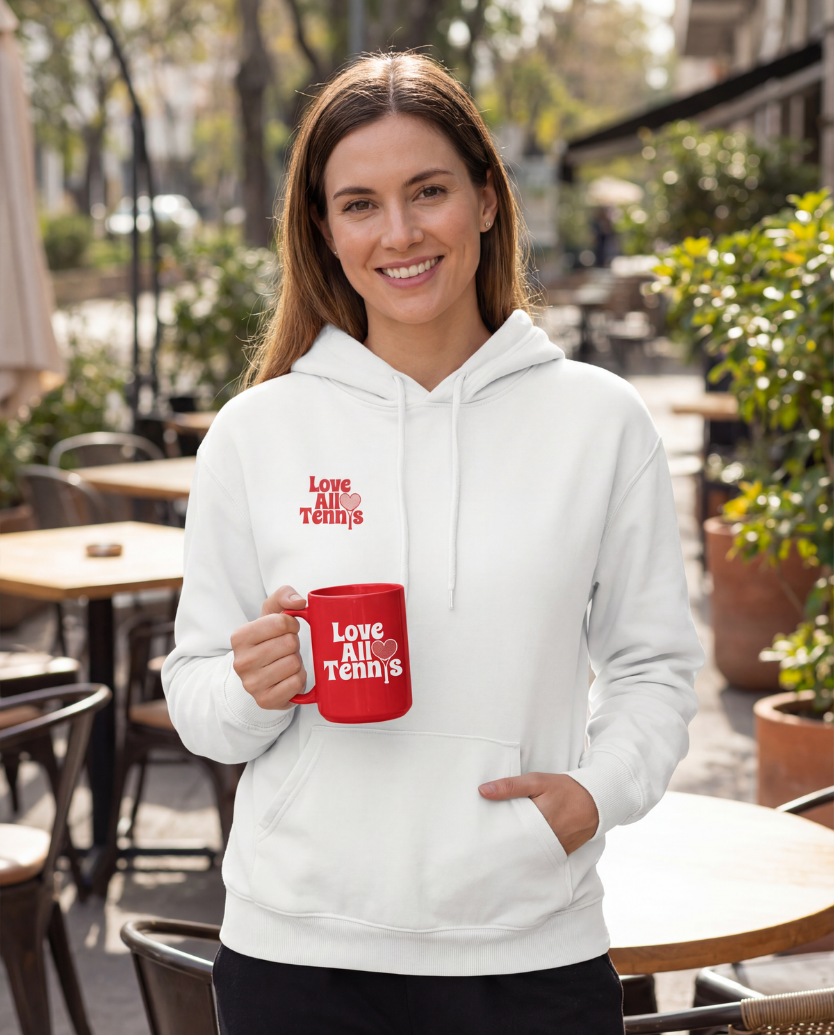 A person in a white sweatshirt holding a bright red ceramic mug, set against a sidewalk of tables