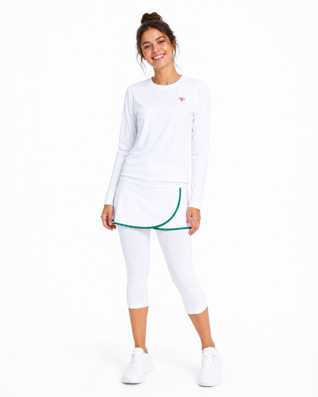 A woman modeling a white long-sleeve athletic top, white capri leggings, and white sneakers against a plain white background. The outfit features a small red logo on the chest and a green curved trim detail along the hem of the top.