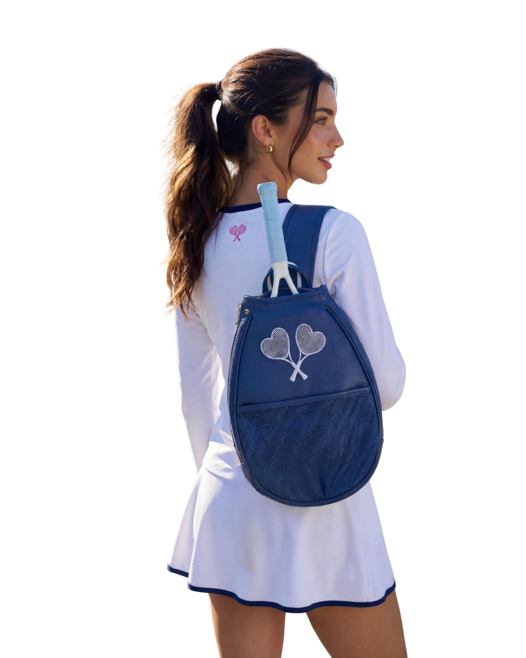 A woman in a white tennis dress with navy trim is shown from the back, wearing a navy blue tennis backpack with a silver heart-and-tie logo and holding tennis rackets on her back, against a solid white background.