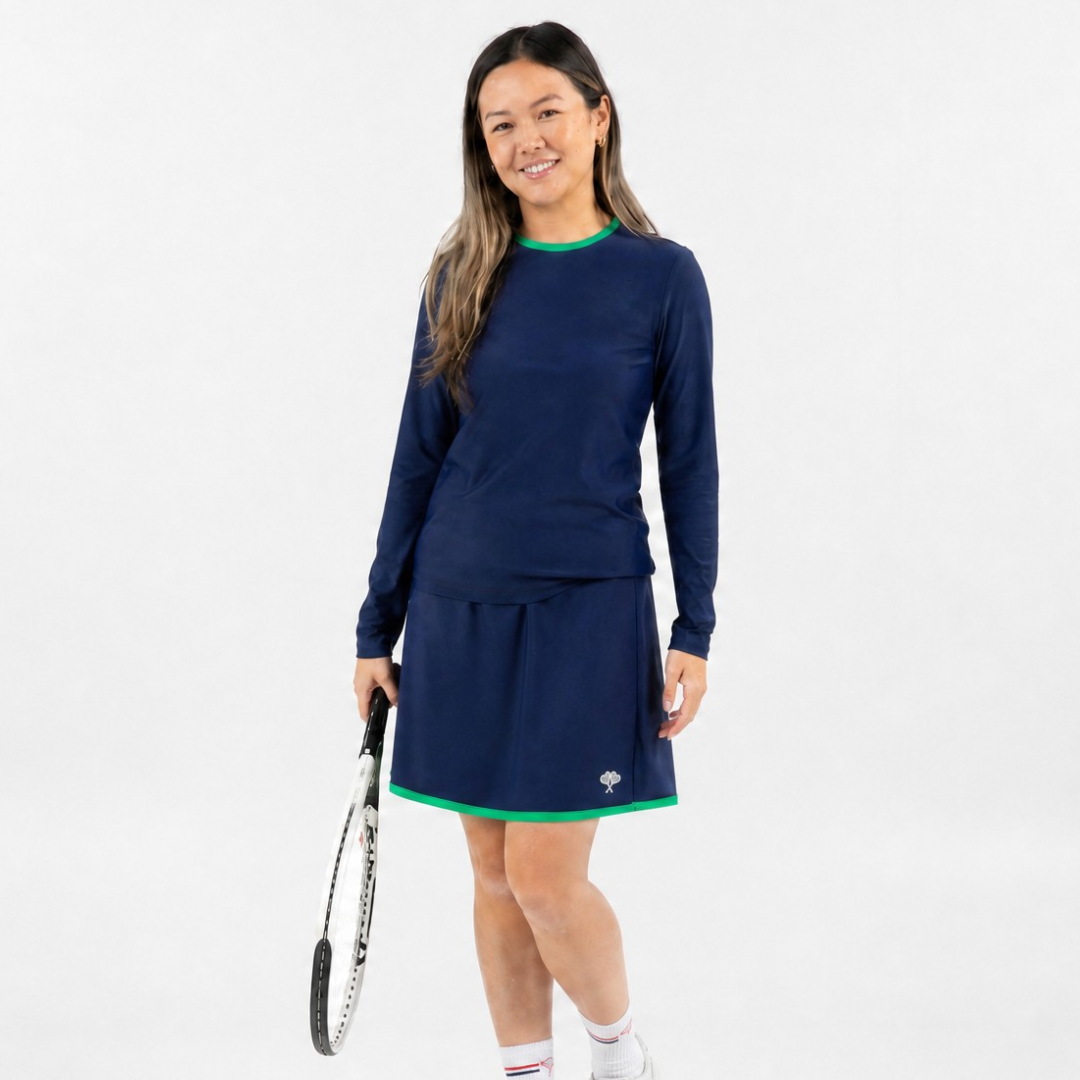 A woman modeling a navy blue long-sleeve tennis dress with green trim, holding a tennis racket against a plain white studio background.