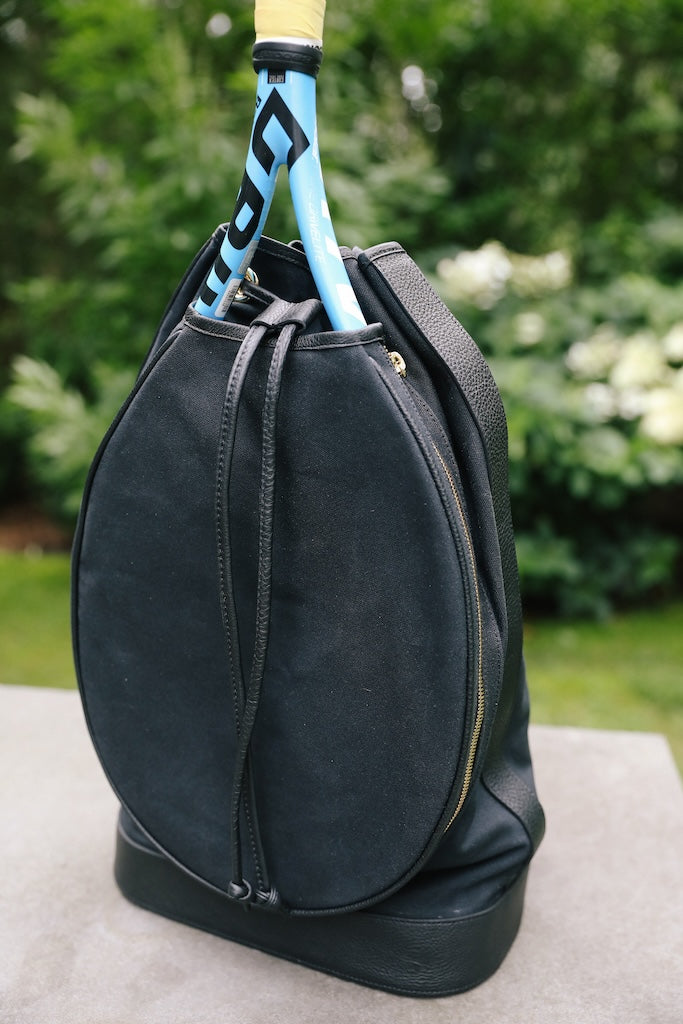 A black tennis bag with a drawstring closure and a zippered base, shown outdoors with a tennis racket tucked inside. The background is a blurred green garden.