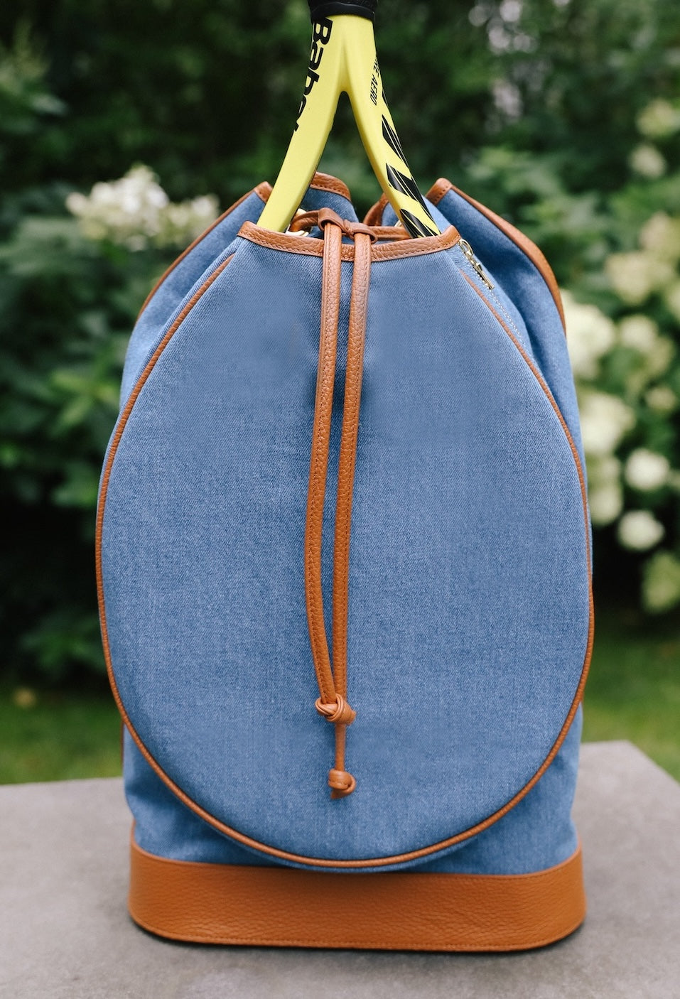 A blue denim-like drawstring backpack with tan leather trim and handles, shown outdoors on a stone surface with green foliage blurred in the background.