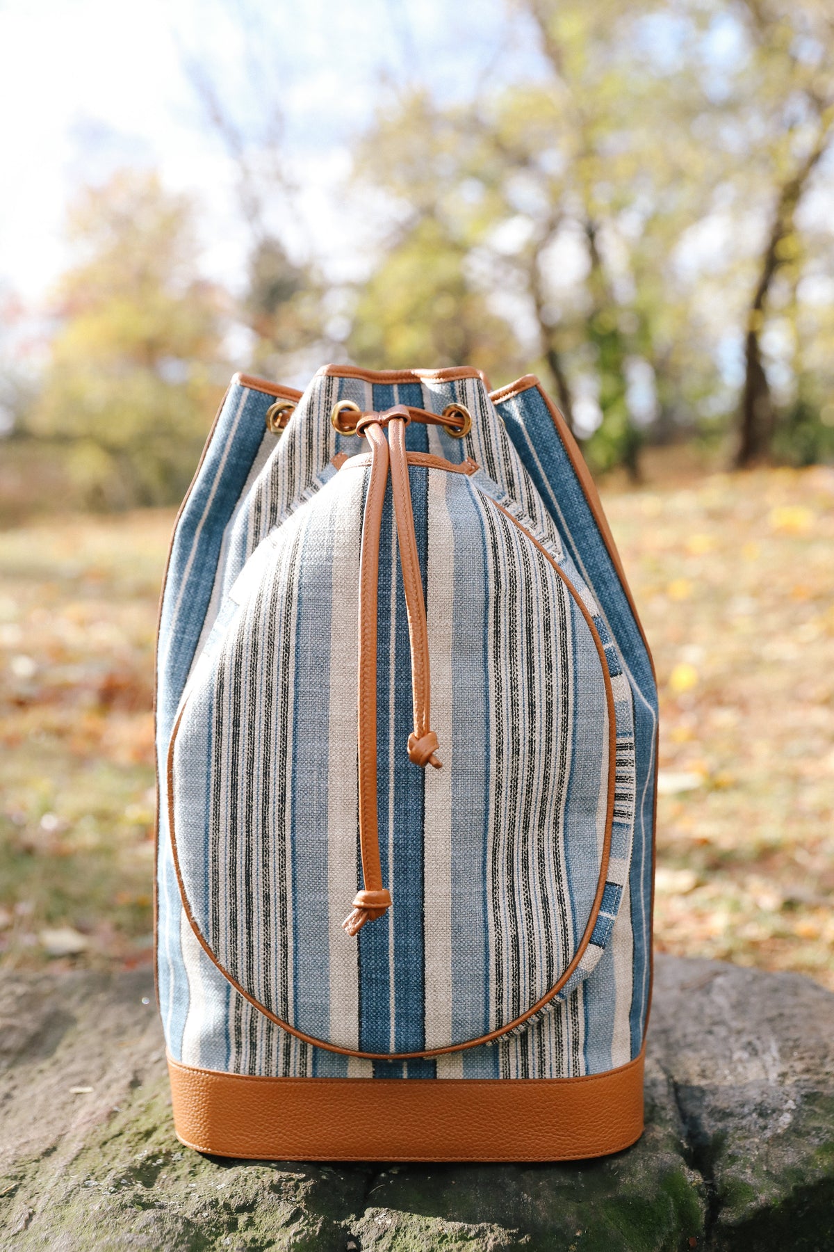 A striped canvas bucket bag with tan leather trim and drawstring closure, photographed outdoors on a stone surface with a blurred autumn park background.