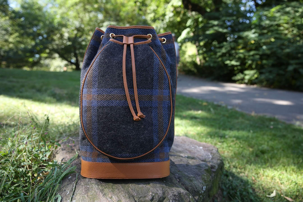 A blue and gray plaid bucket bag with tan leather trim and drawstring, standing on a stone in a sunny park setting.