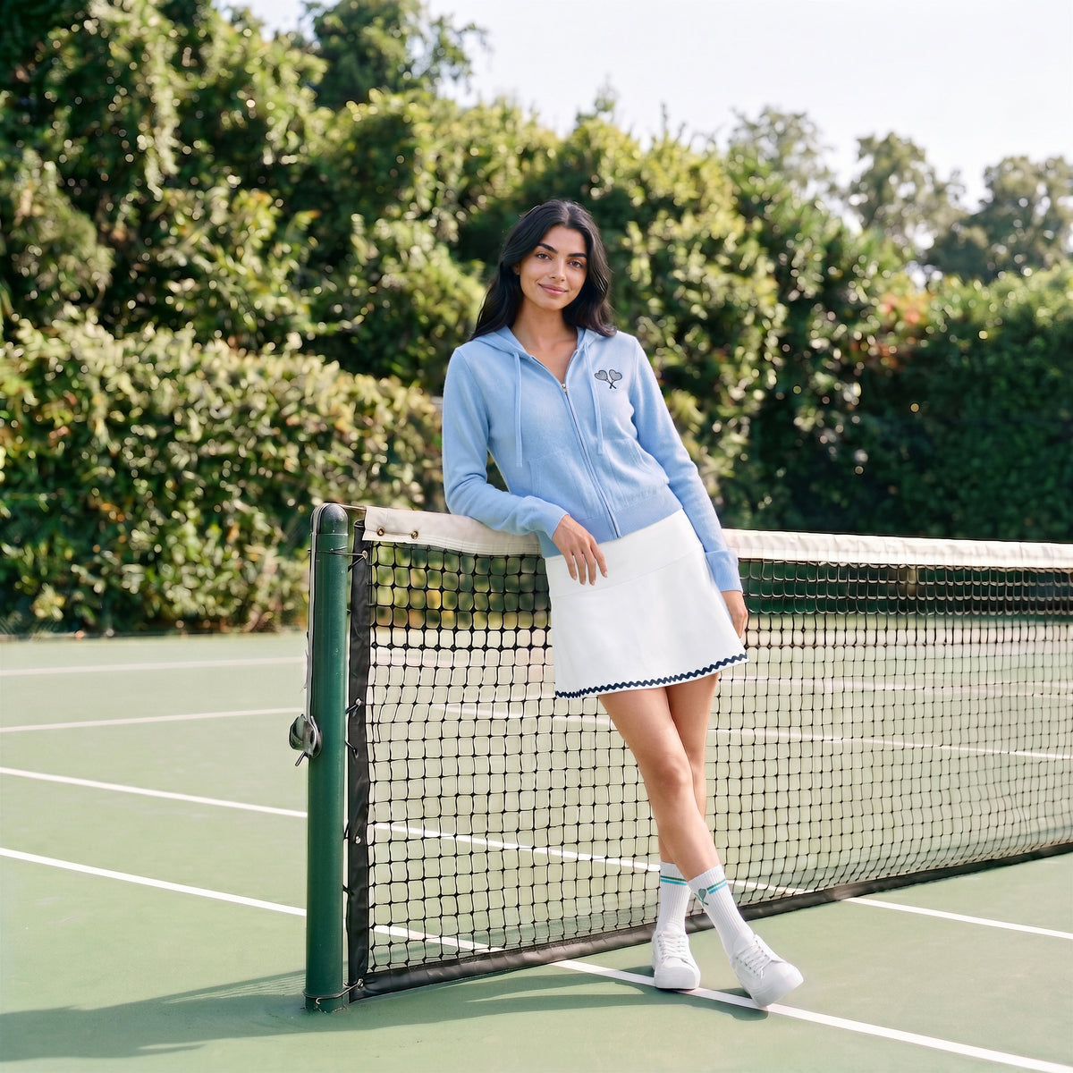 A woman stands on a tennis court leaning against the net, wearing a light blue zip-up hoodie with a small embroidered logo on the chest, a white tennis skirt, white socks, and white sneakers.