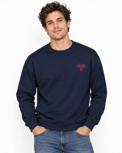 A man wearing a navy blue crewneck sweatshirt with a small red embroidered heart and crossed keys motif on the left chest, paired with blue jeans, against a plain white background.