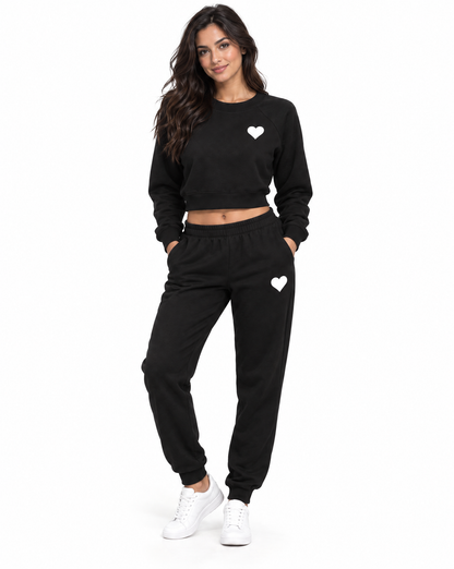 A woman modeling a black two-piece sweatshirt and jogger set with small white heart graphics on the chest and thigh, paired with white sneakers, against a plain white background.