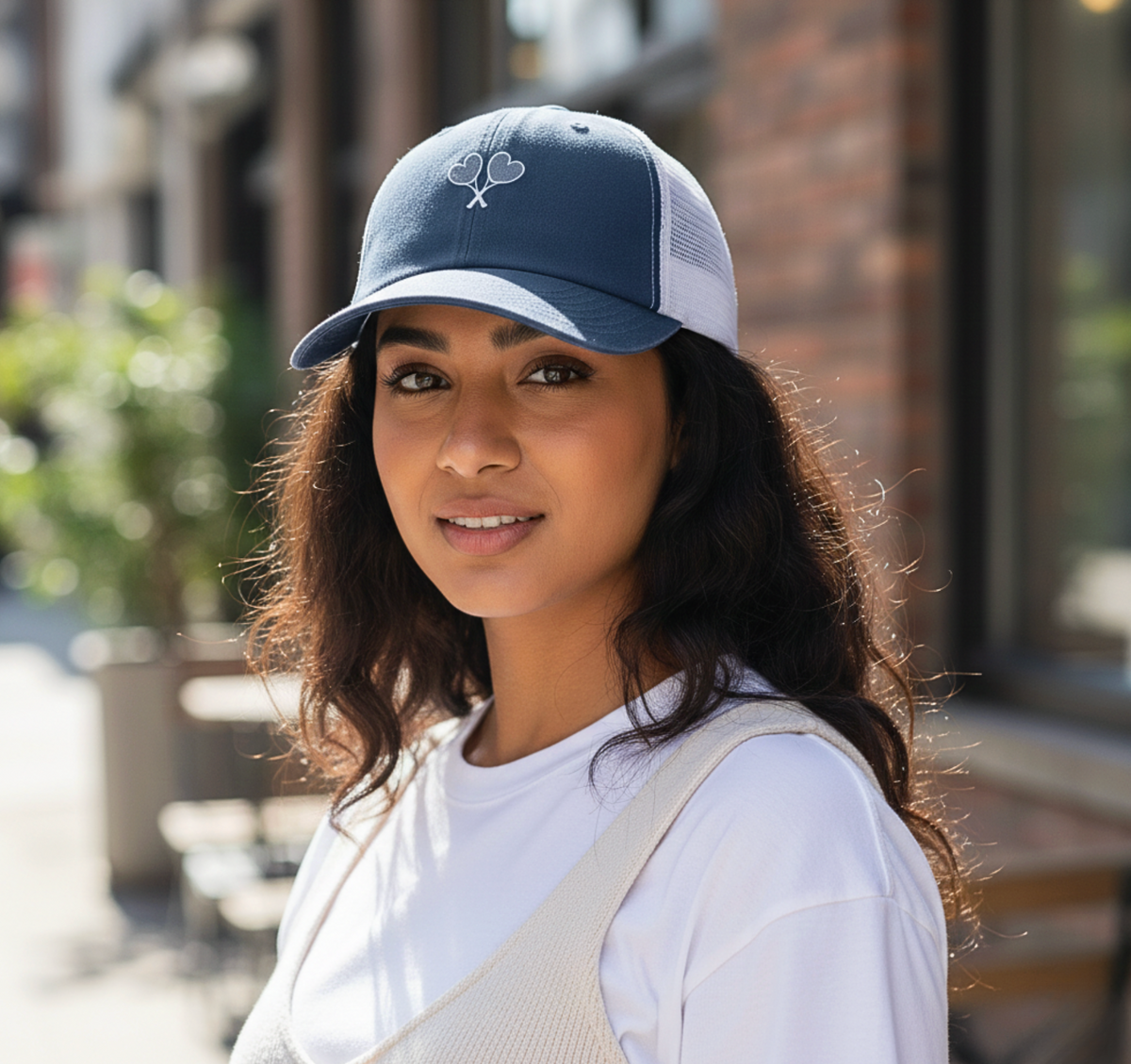 Woman wearing a blue and white trucker hat with heart shaped tennis rackets crossing on the front 