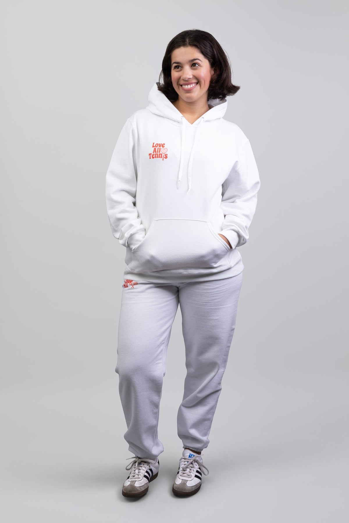 Woman wearing white sweatsuit with red logos and sneakers set against a grey studio background