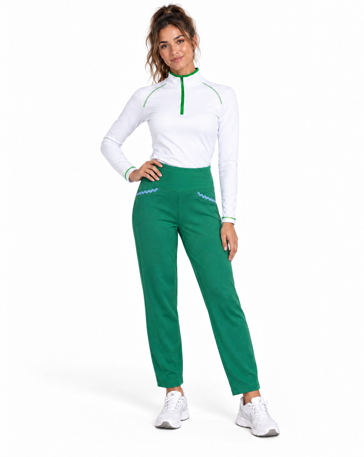 A woman modeling a white long-sleeve athletic top with green trim and green high-waisted pants, paired with white sneakers, against a plain white background.