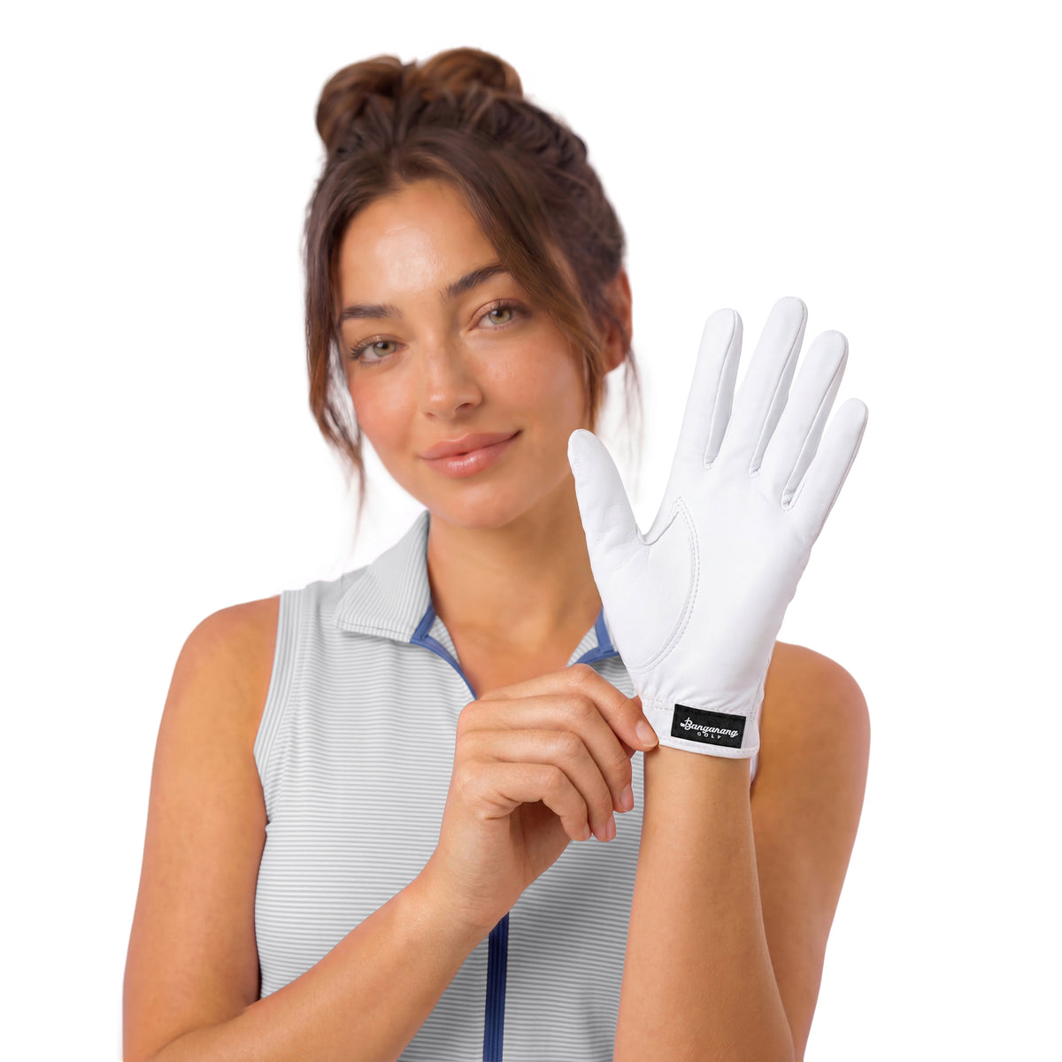 A woman wearing a light gray sleeveless top holds up a white golf glove with a black logo patch on the cuff, set against a solid white background.