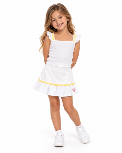 A young girl with long light brown hair stands against a white studio background wearing a white sleeveless top and skirt with yellow trim and white sneakers.