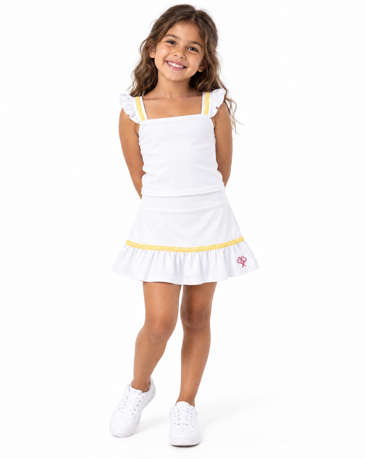 A young girl with long brown hair is smiling and posing in a white sleeveless tennis-style dress with yellow patterned trim and white sneakers, photographed against a seamless white background.