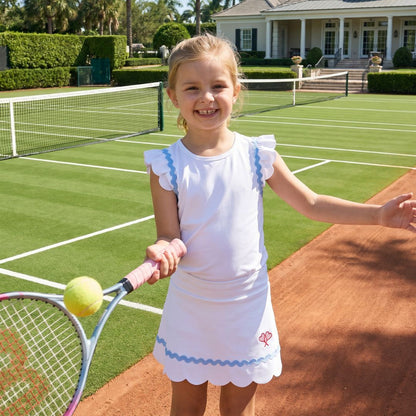 Young girl on a tennis court holding a racket and ball, with a house and palm trees in the background.