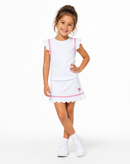 A young girl modeling a white two-piece dress set with pink scalloped trim details, consisting of a short-sleeve top and a matching skirt, paired with white socks and sneakers against a plain white background.