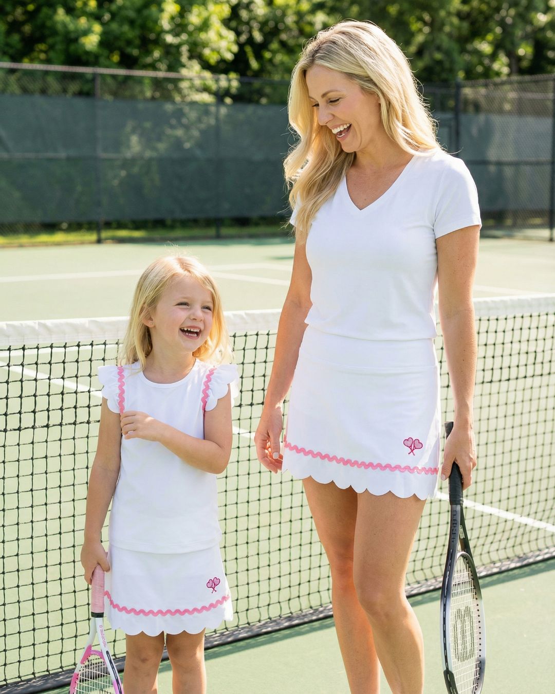 Woman and young girl on a tennis court wearing matching white outfits with pink trim.
