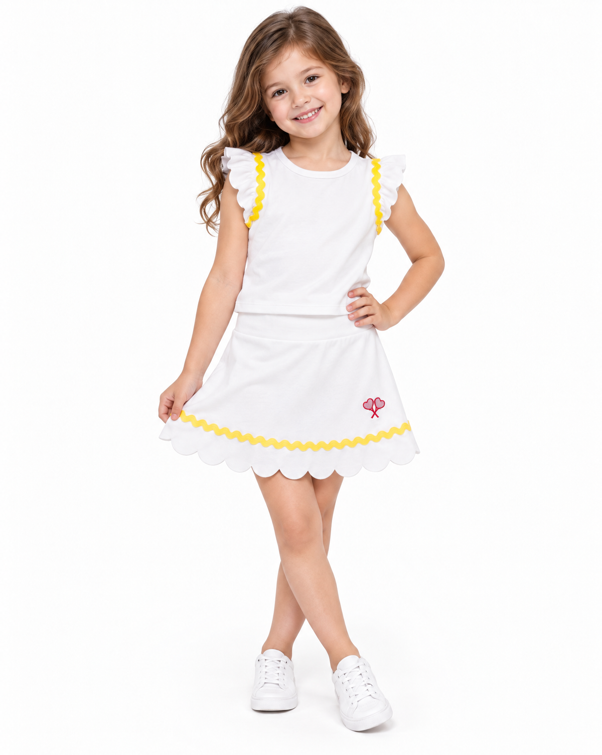 A young girl with long brown hair stands against a white studio background wearing a white sleeveless dress with yellow ruffle trim, a yellow scalloped hem, and a small red logo, paired with white sneakers.