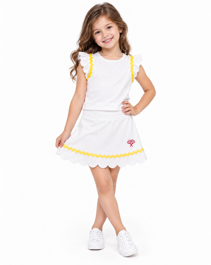 A young girl with long brown hair stands against a white studio background wearing a white sleeveless dress with yellow ruffle trim, a yellow scalloped hem, and a small red logo, paired with white sneakers.