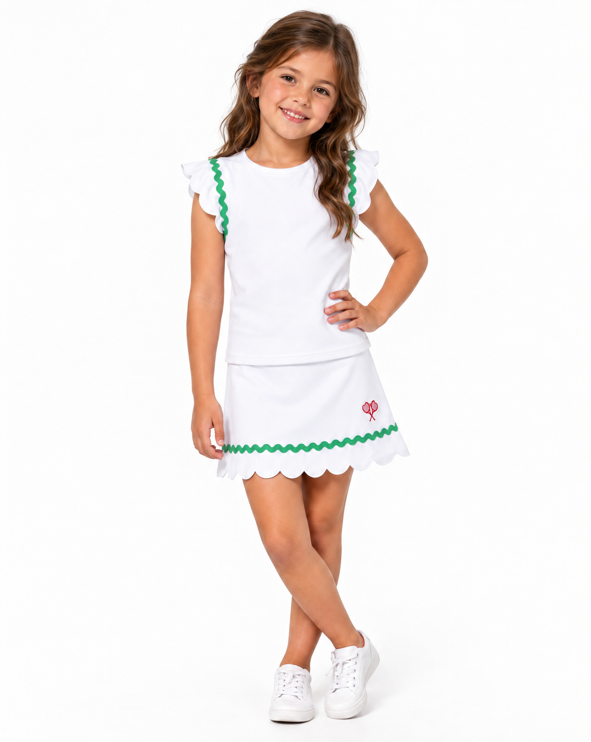 A young girl with long brown hair stands against a white studio background wearing a white sleeveless top and skirt with green scalloped trim and a small red logo, paired with white sneakers.