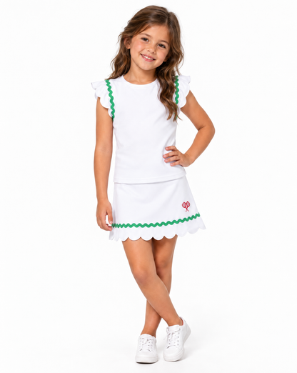 A young girl with long brown hair stands against a white studio background wearing a white sleeveless top and skirt with green scalloped trim and a small red logo, paired with white sneakers.