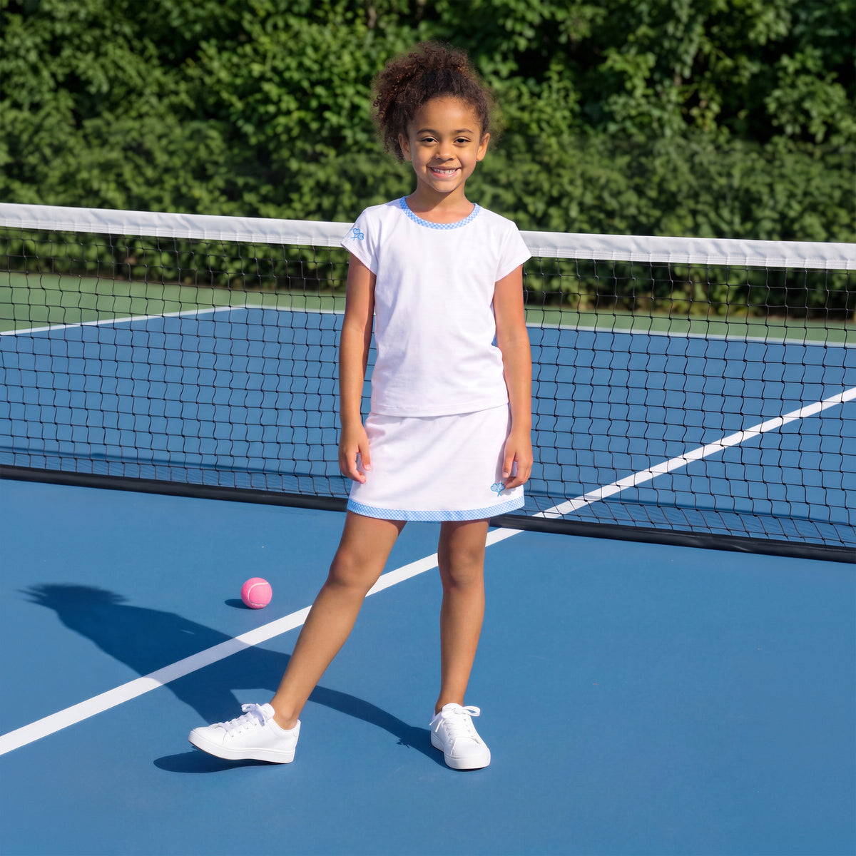 A young girl stands on a blue tennis court wearing a white short-sleeve tennis top and skirt with light blue trim. A pink tennis ball lies on the court near her shadow, with a net in front of her and green trees in the background.