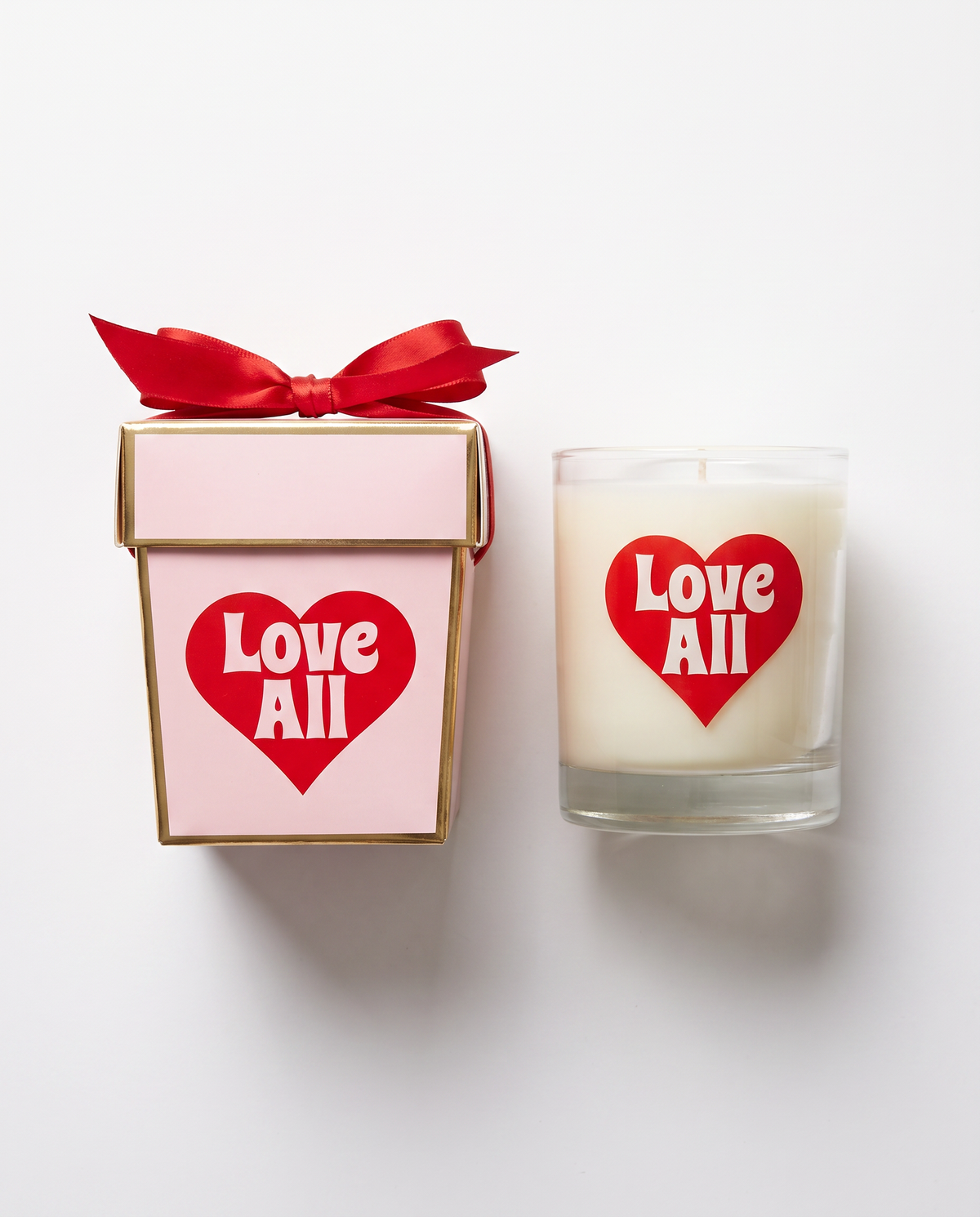 A white candle in a clear glass jar with a red heart label reading 'Love All' sits beside a pink gift box with a red ribbon and matching heart label on a white background.