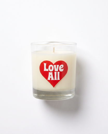 A white wax candle in a clear glass jar with a red heart-shaped sticker on the front that reads 'Love All' in white text, photographed against a plain white background.