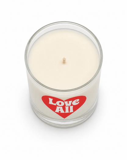 An off-white wax candle in a clear glass jar with a red heart-shaped label that reads 'Love All' in white text, set against a plain white background.