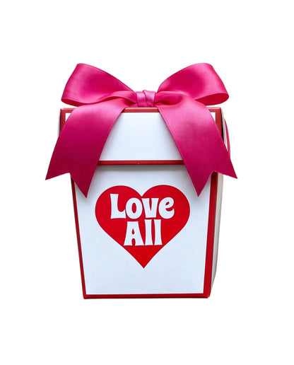 A white gift box with red trim, tied with a large pink satin bow, featuring a red heart on the front with the words 'Love All' in white.