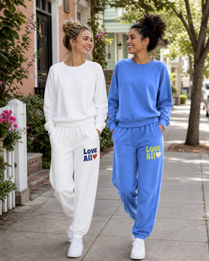 Two women walking side-by-side on a sidewalk wearing matching sweatshirt and jogger sets. The woman on the left wears a white set with 'Love All' printed in blue and red on the left leg.