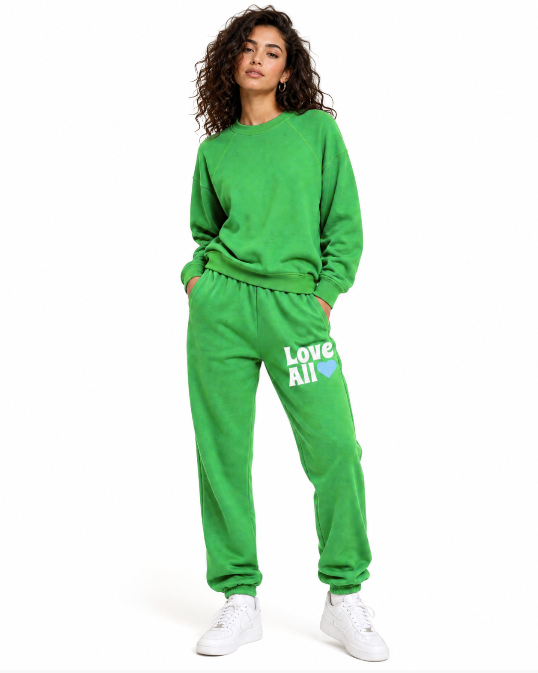 A woman modeling a bright green sweatshirt and matching sweatpants set. The sweatpants feature the text 'Love All' in white with a small blue heart on the left thigh.