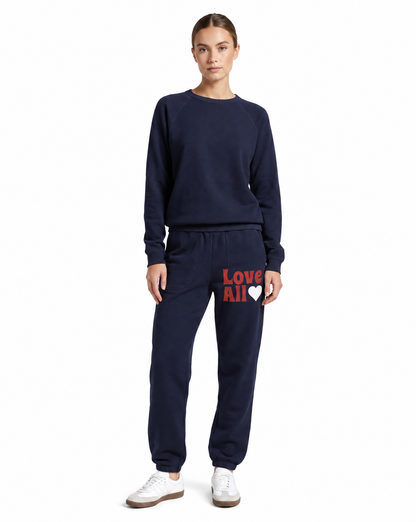 A woman modeling a navy blue sweatshirt and matching jogger pants set. The jogger pants feature the text 'Love All' in red and a white heart on the left thigh.