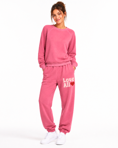 A woman modeling a matching pink sweatshirt and jogger pants set with white sneakers, standing against a plain white studio background. The sweatshirt has a crew neck and long sleeves, while the joggers feature a white 'Love All' text and red heart graphi