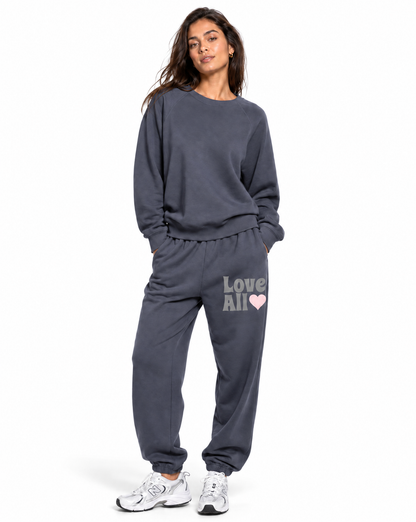 A woman modeling a dark gray sweatshirt and jogger sweatpants set. The sweatpants feature the text 'Love All' in white with a small pink heart on the left thigh.